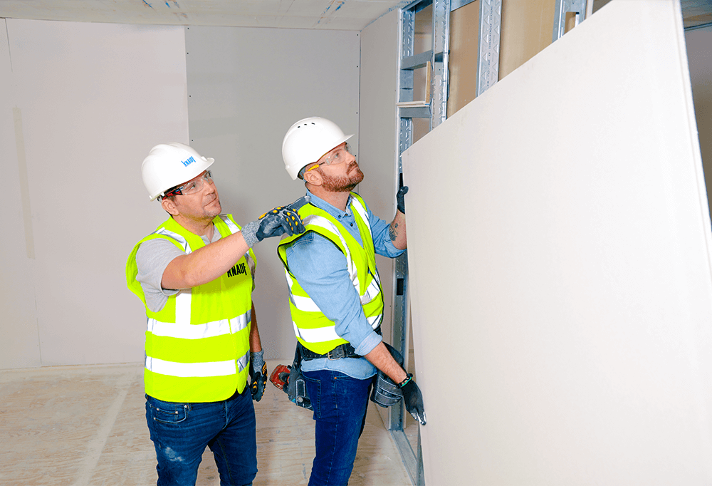 A Knauf employee in a high vis guides a customer with a high vis on how to install plasterboard onto a metal frame