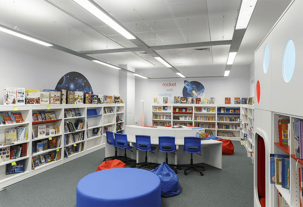 A modern school library with books on shelves surrounding the walls and a row of chairs around a desk in the middle