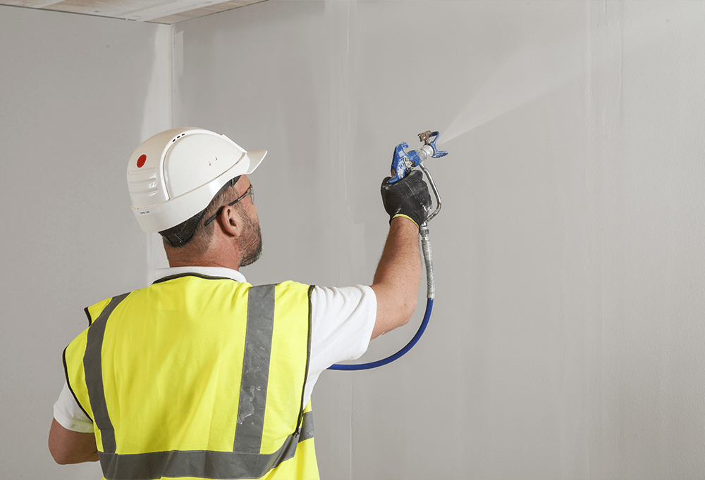 A Knauf trainer in a high vis is using a spray machine onto a wall in an indoor construction site