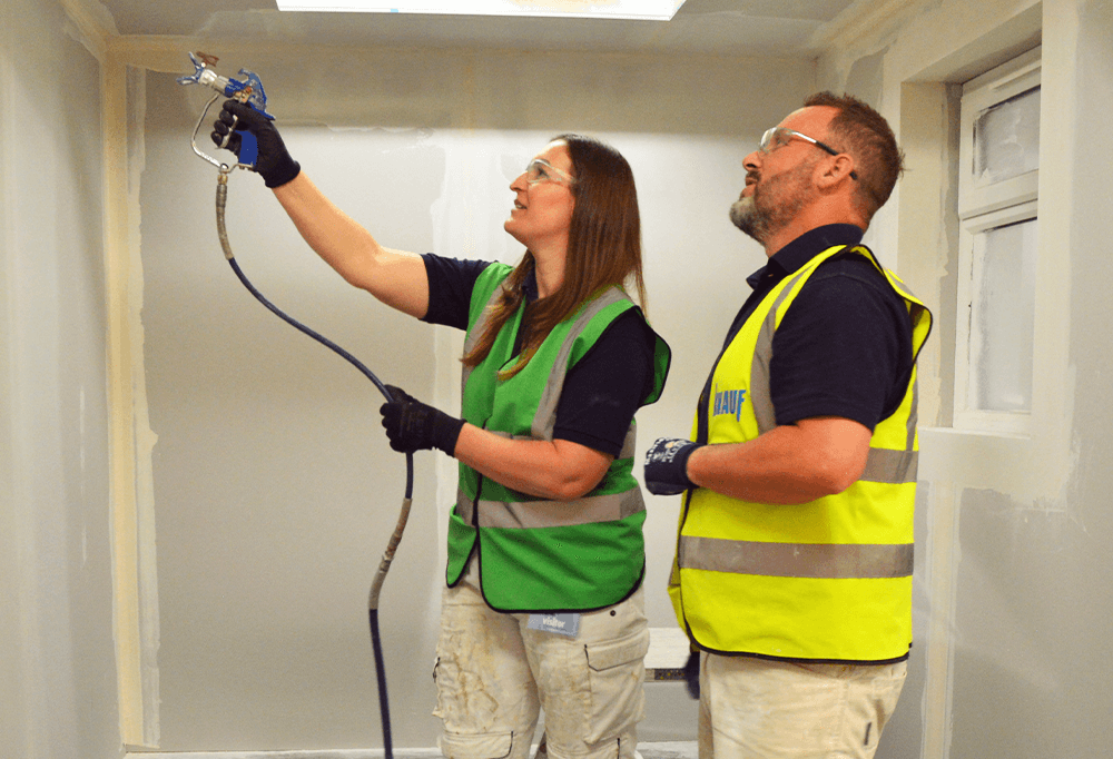 A male Knauf trainer in a high vis shows a female customer how to use a spray plaster machine in an indoor construction site