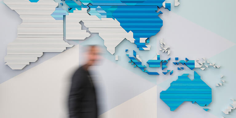 A man, blurred to show they are walking, moves past a modern style world map mounted to the wall in blue and white